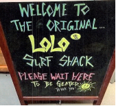 A sandwich-board chalk sign with colourful handwritten text that reads: "Welcome to the original Lolo Surf Shack. Please wait here to be seated. Thank you." The word Lolo appears in a different colour and font followed immediately by a trademark registration symbol.
